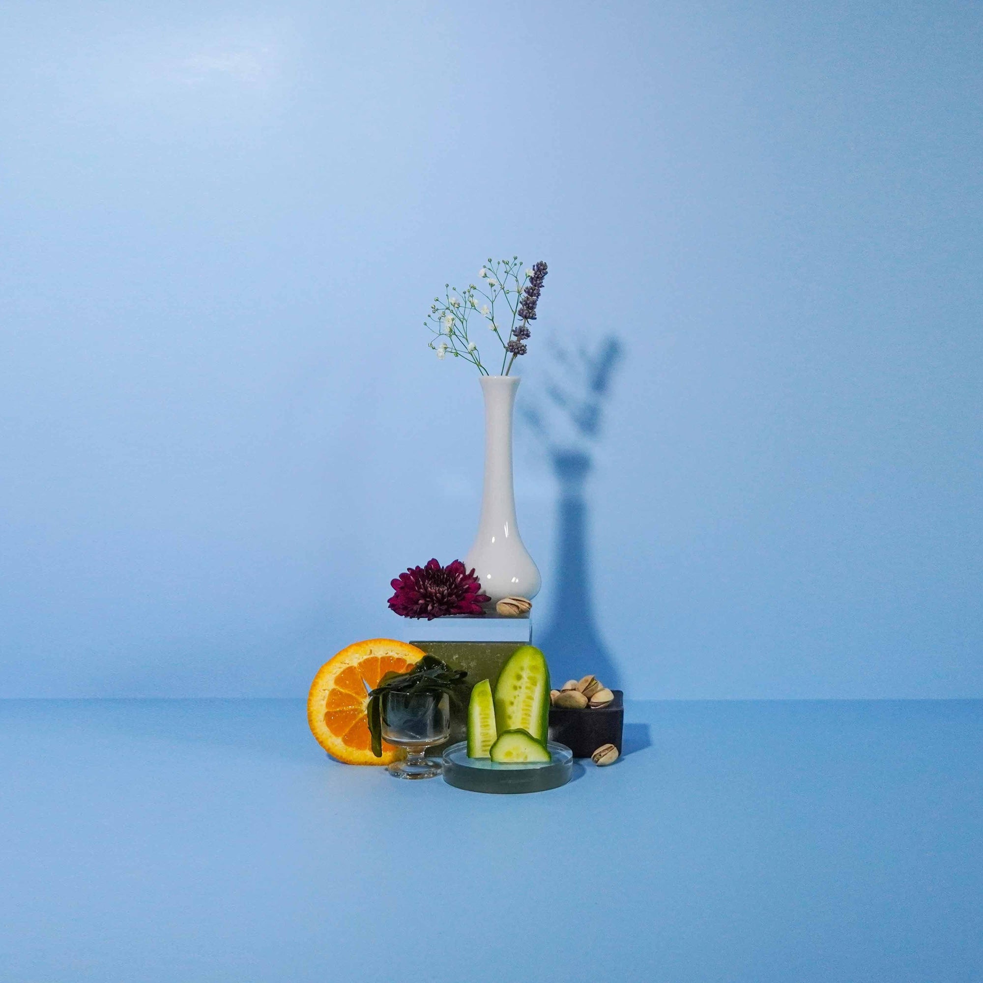 A bottle of fragrance oil labeled 'Spanish Orange + Cucumber Fizz' with a visual representation of orange slices and cucumber pieces in front of the bottle, set against a blue background.