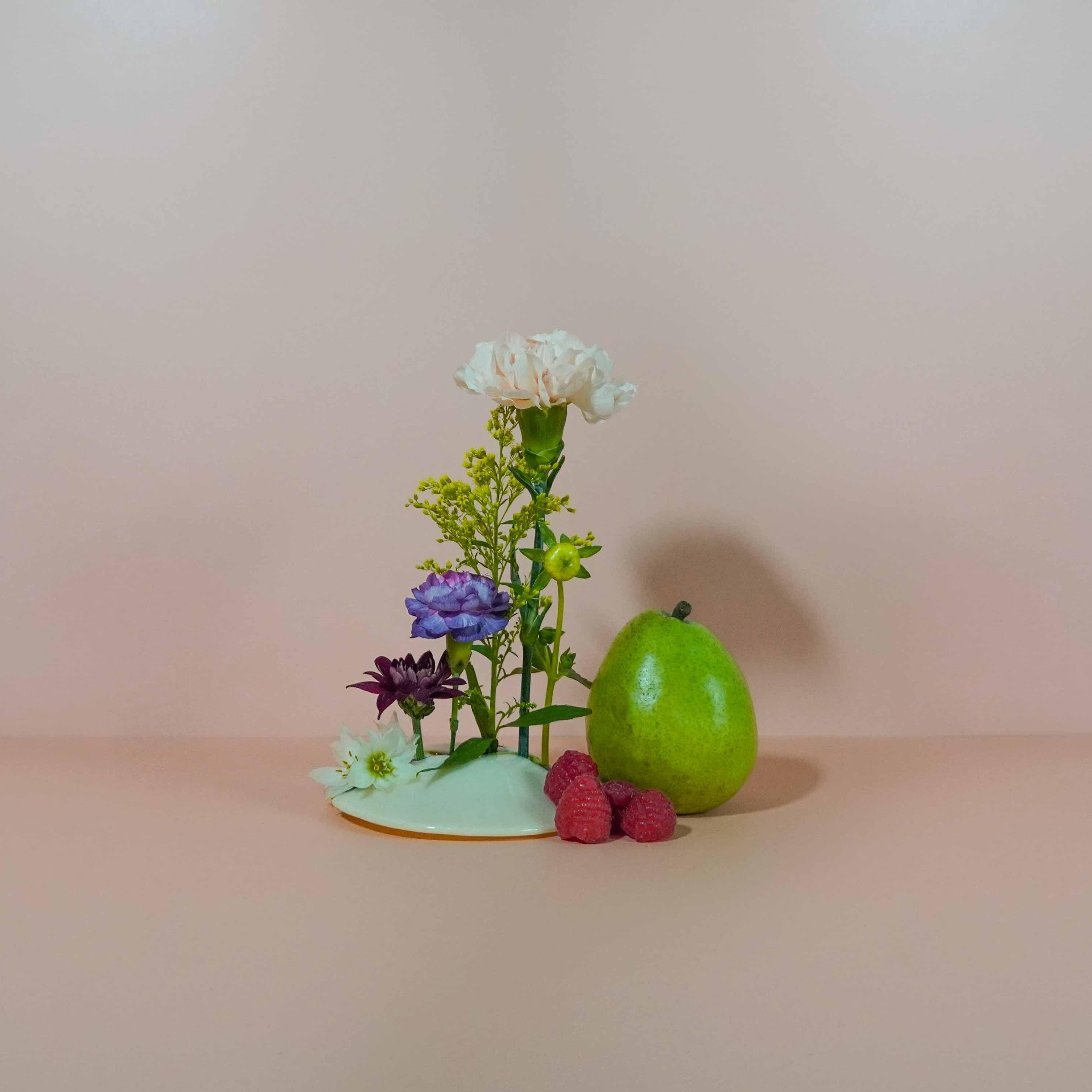 A small decorative item featuring a green apple, raspberries, and artificial flowers arranged on a pastel background.