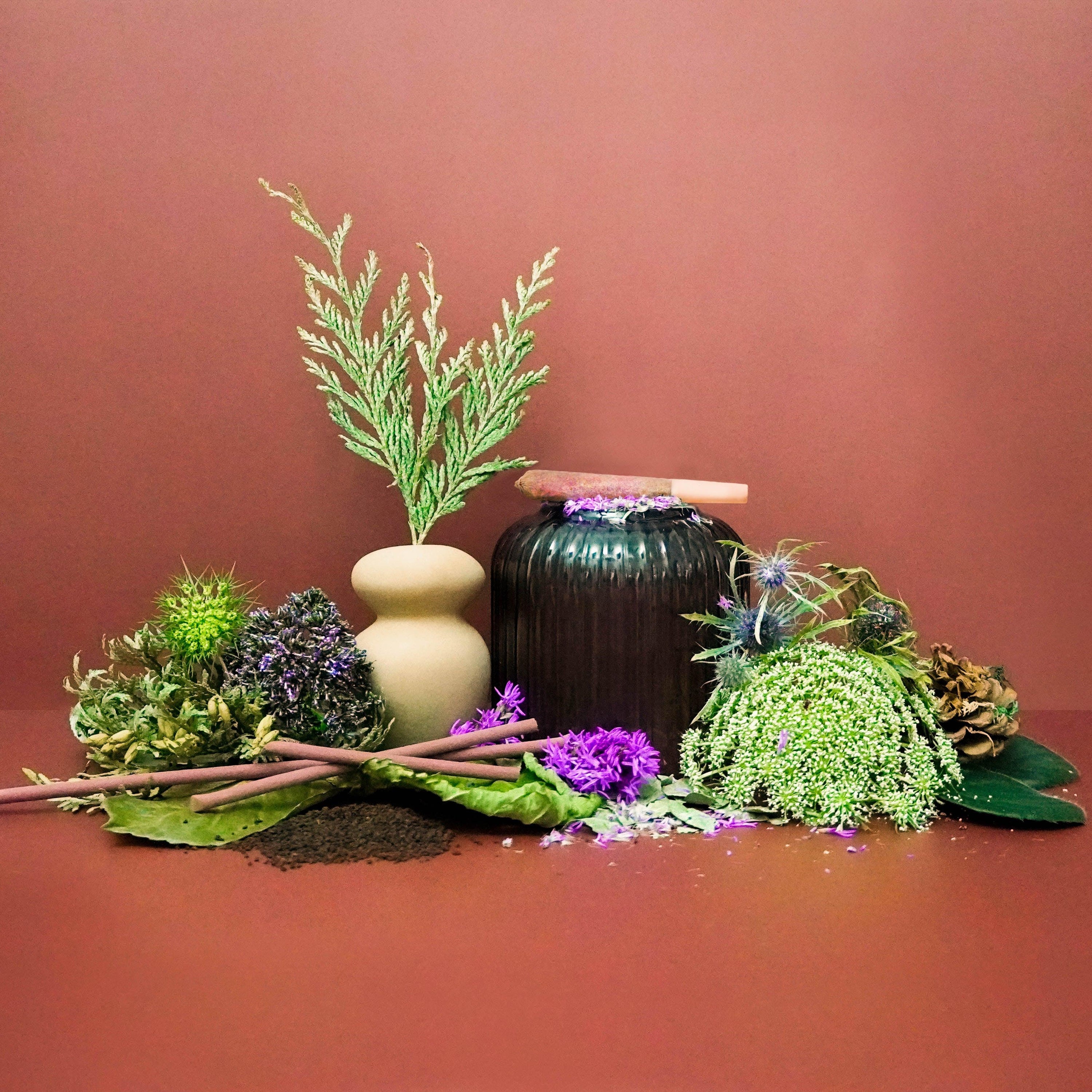 A still life setup featuring a variety of green plants and flowers, with a few beige and black decorative objects, presented against a brown background.