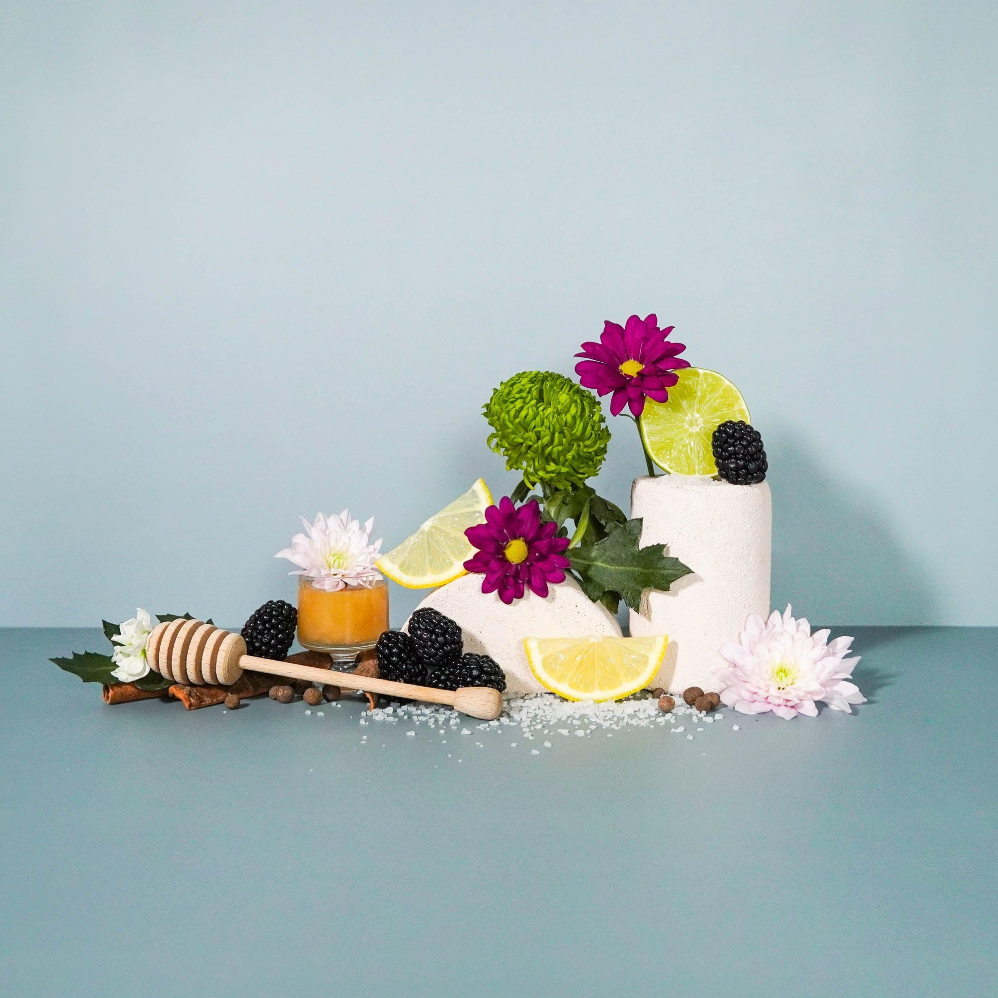 A still life arrangement featuring a white cup with blackberry tea, surrounded by lemon and lime slices, a honey dipper, and a variety of decorative flowers against a blue background.